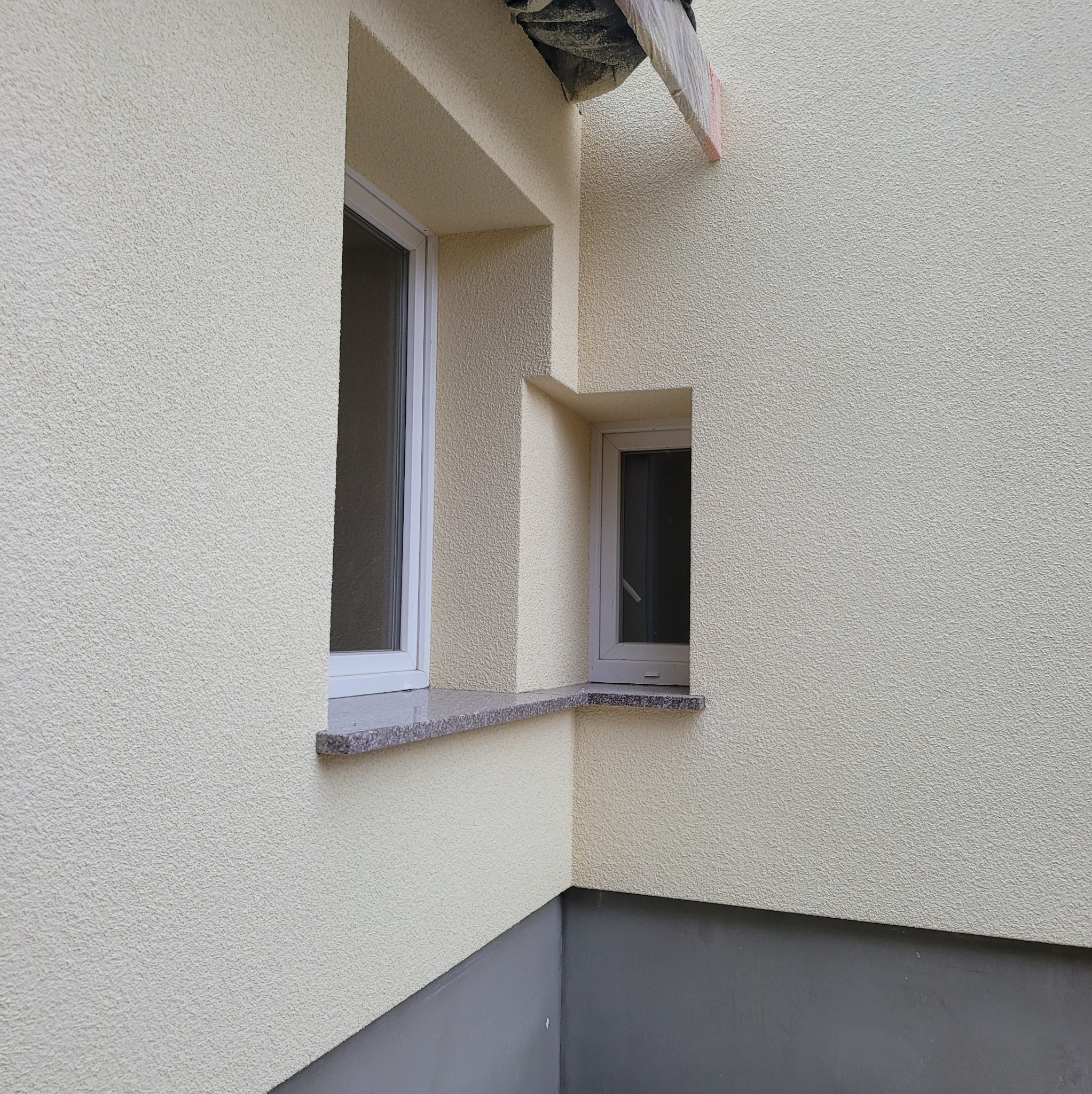 Fragment elewacji budynku z dwoma oknami o różnej wielkości, wykończonej jasnym tynkiem strukturalnym i ciemniejszą podmurówką.