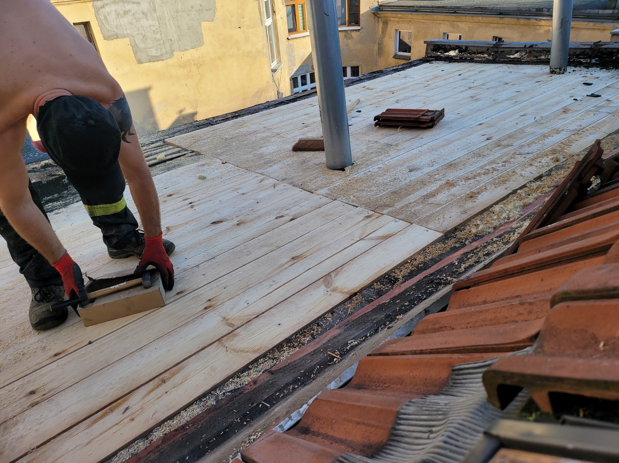 Pracownik w czerwonych rękawicach montuje drewniane deski na dachu, widoczne narzędzia i fragment starej dachówki.