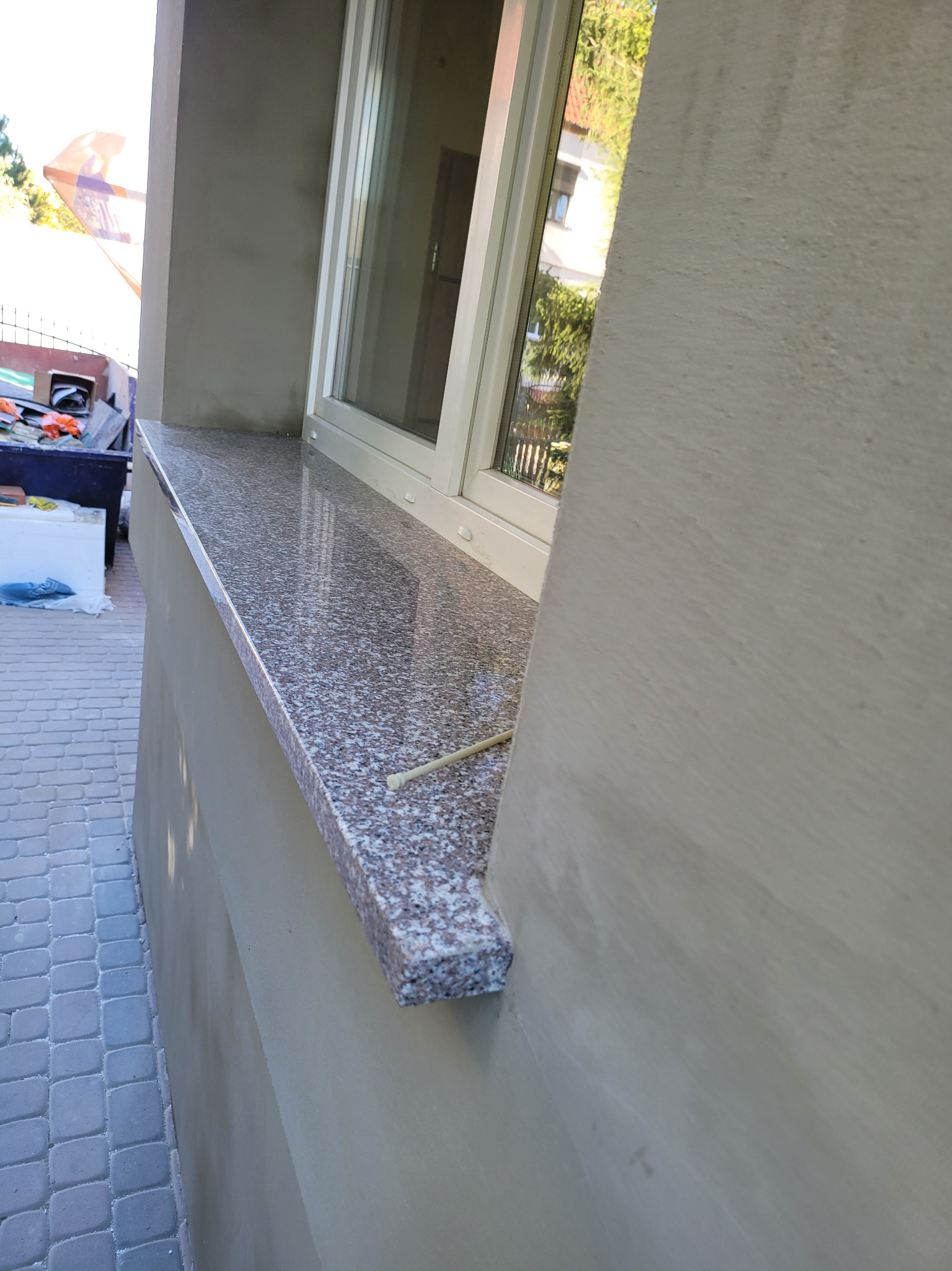 Zewnętrzny parapet z szarego granitu z widoczną fakturą, zamontowany pod oknem w budynku z nową elewacją, na pierwszym planie fragment chodnika z kostki brukowej.