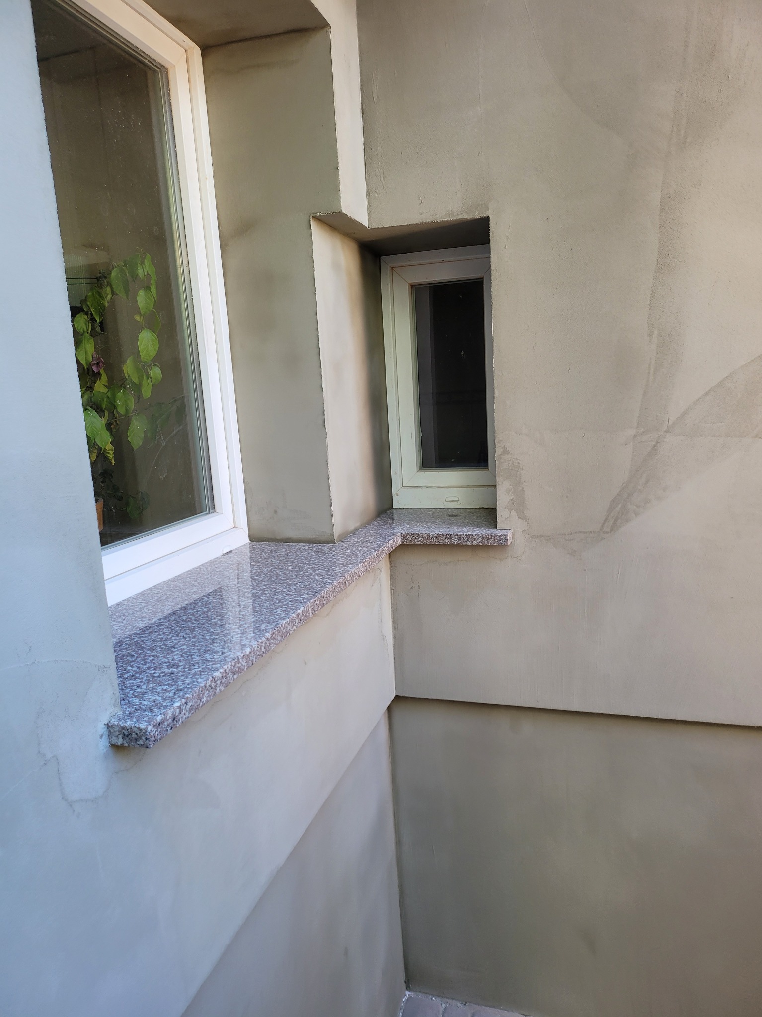 Ściana zewnętrzna budynku po nałożeniu szarego tynku cementowo-wapiennego, z dwoma oknami o różnych rozmiarach i granitowymi parapetami o szarej barwie z różowymi plamkami.