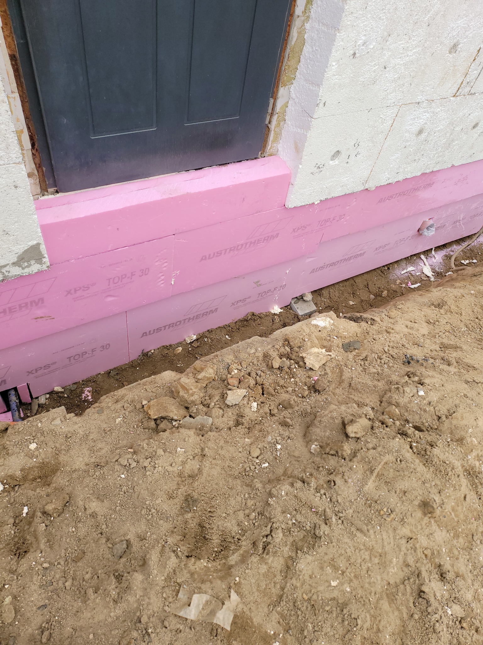 Ocieplenie fundamentu różowym styrodurem Austrotherm XPS TOP-F 30, widoczne fragmenty ściany i gruntu. Prace izolacyjne w toku, detale montażu.