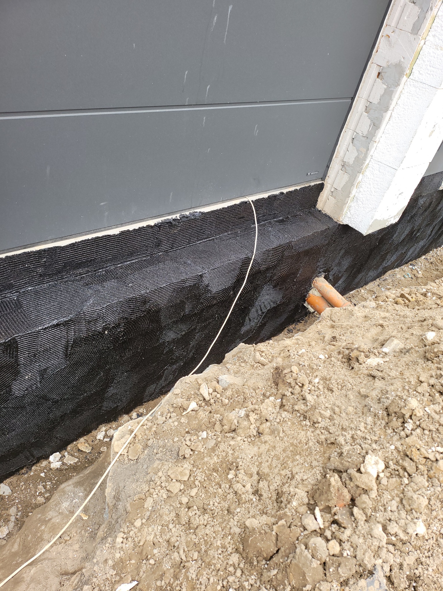 Izolacja fundamentów budynku z widocznymi rurami kanalizacyjnymi i fragmentem ściany z betonu komórkowego. Czarna warstwa izolacyjna na fundamencie.