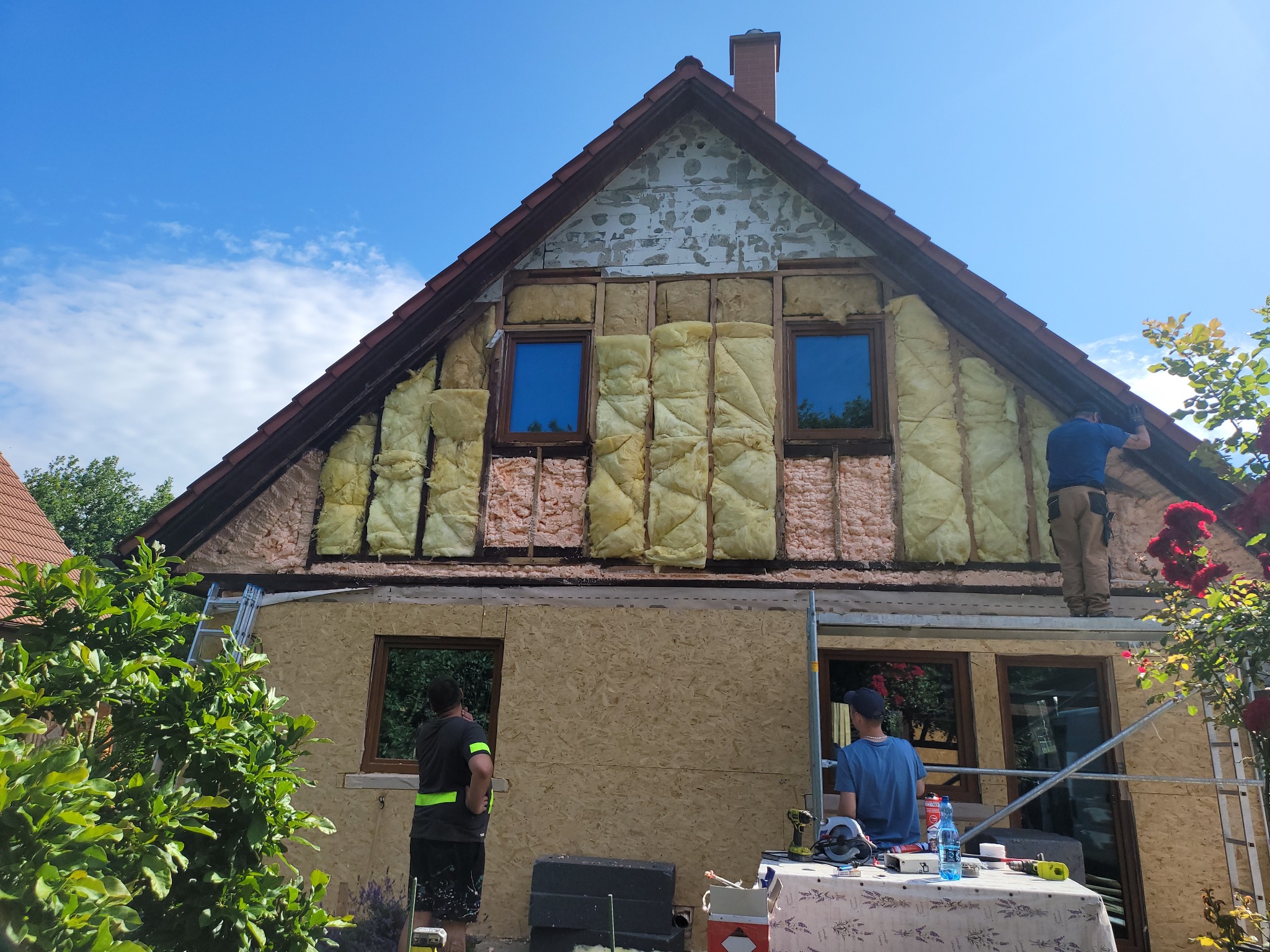 Ocieplanie domu z zewnątrz: pracownicy na rusztowaniu montują izolację i płyty OSB. Widoczne okna, fragment dachu i niebieskie niebo. Prace w toku, dzień słoneczny.