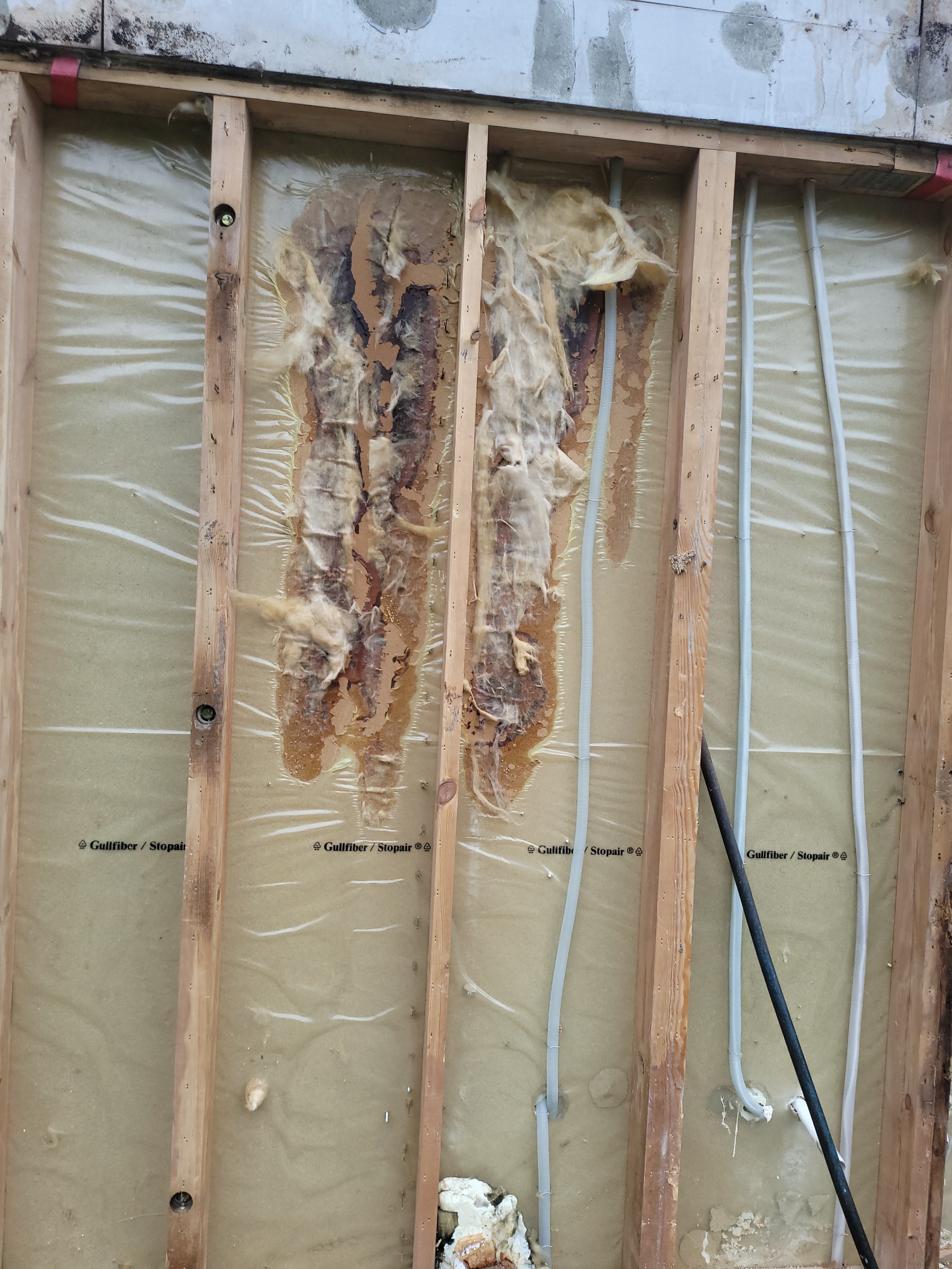 Odsłonięta ściana z drewnianą konstrukcją, izolacją pokrytą folią, z widocznymi uszkodzeniami i instalacjami. Widoczne ślady zawilgocenia na izolacji.