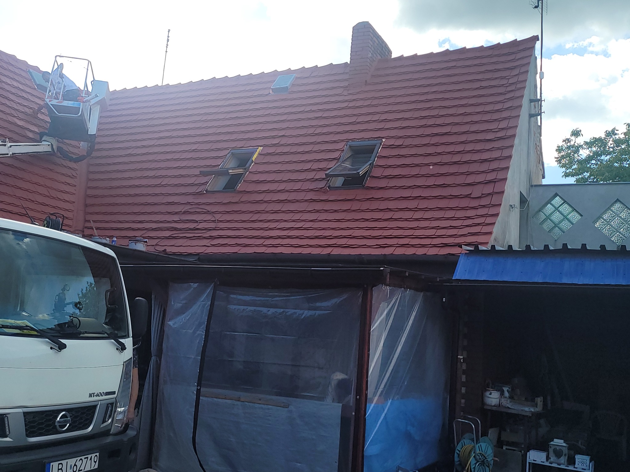 Czerwony dach domu z oknami dachowymi, widziany z dołu. Na dachu pracownik w podnośniku koszowym. Częściowo widoczny biały samochód dostawczy.