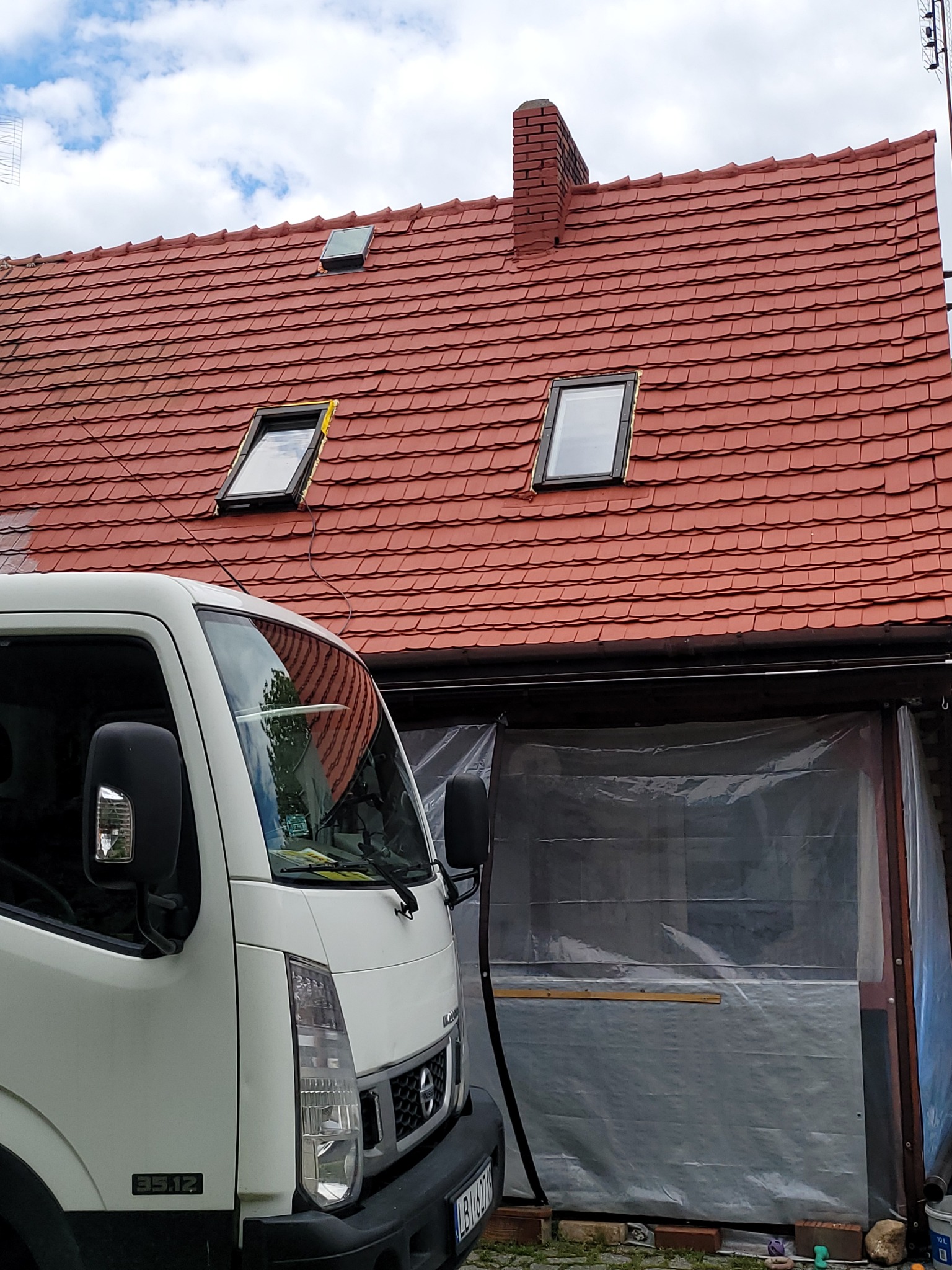 Czerwony dach z dachówką karpiówką i oknami dachowymi, widoczny fragment białego samochodu dostawczego i zadaszone miejsce pracy, zabezpieczone folią.