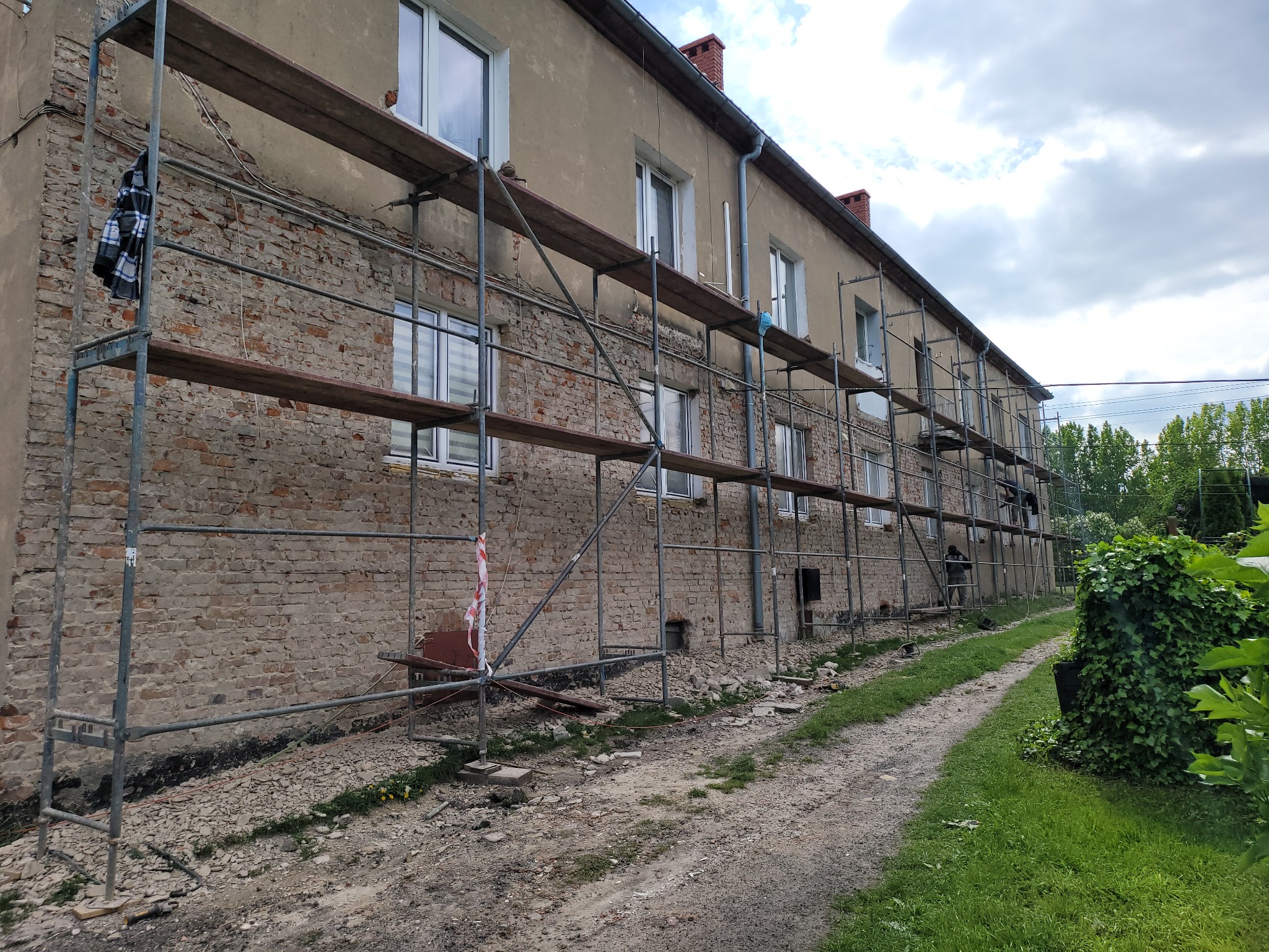 Budynek w trakcie renowacji elewacji z odsłoniętą cegłą i rusztowaniami. Widoczne okna z białymi ramami i fragment drogi gruntowej.