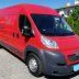 Czerwony samochód dostawczy marki Peugeot z niebieską tablicą rejestracyjną, zaparkowany przed budynkiem z zielonymi krzewami w tle, sfotografowany w słoneczny dzień.