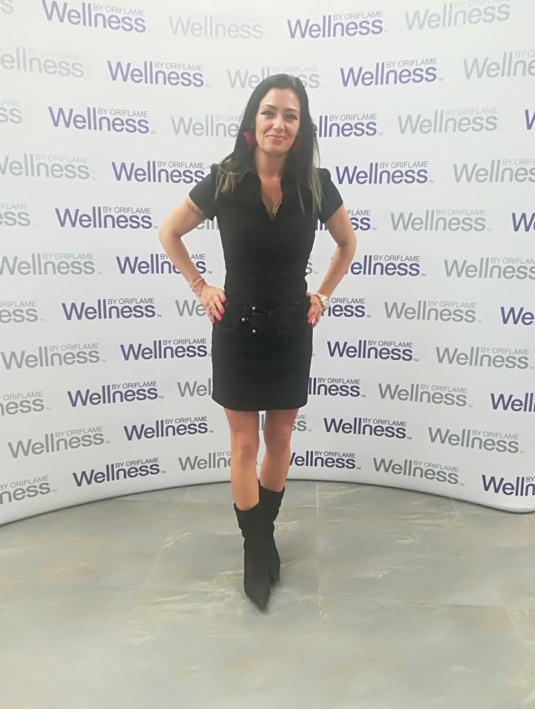 Kobieta w czarnej sukience i kozakach pozuje na tle ściany z logo Wellness by Oriflame, prezentując makijaż i stylizację.