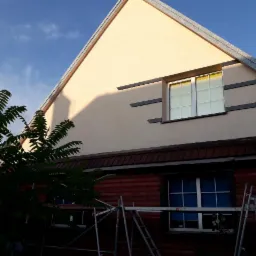 Wykończona elewacja domu z beżowym tynkiem, ozdobnymi czarnymi pasami i oknem, częściowo zasłonięta przez rusztowanie i zieleń drzewa.