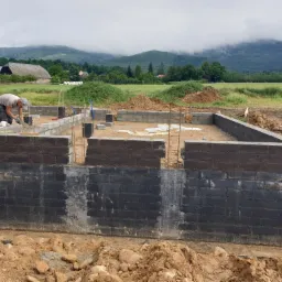 Budowa fundamentów z bloczków betonowych, zabezpieczonych czarną izolacją pionową, w tle krajobraz z budynkami gospodarczymi i zalesionymi wzgórzami pod mglistym niebem.