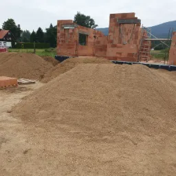 Wznoszone mury domu z czerwonej cegły ceramicznej Porotherm na fundamencie, z widocznymi stertami piasku na placu budowy i rusztowaniem w tle, w oddali zabudowania wiejskie i zalesione wzgórza.