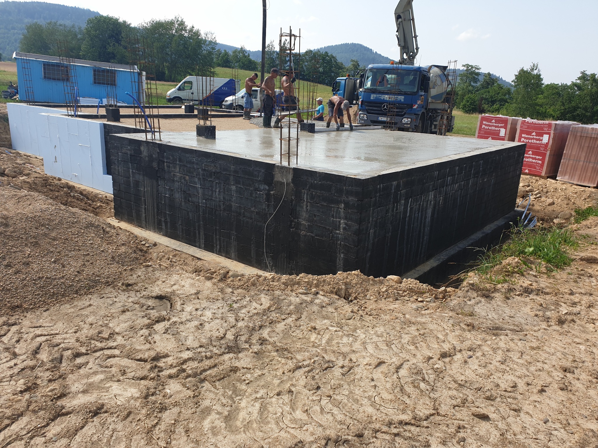 Wylewanie betonu na fundamenty domu jednorodzinnego; widoczni pracownicy, betonowa pompa na ciężarówce, izolacja termiczna fundamentów i zbrojenie.