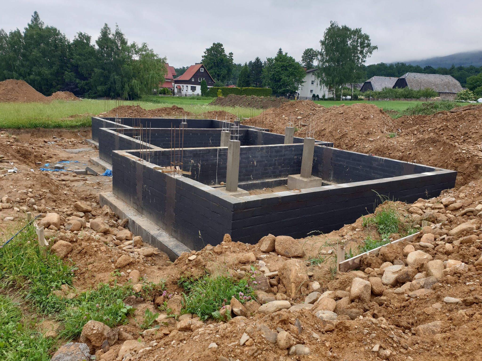 Wylane fundamenty pod budynek z betonu pomalowane na czarno, widoczne zbrojenia i betonowe słupy, otoczone ziemią i kamieniami, w tle zabudowa wiejska i zalesione wzgórza.