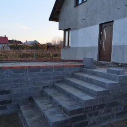 Wznoszone schody wejściowe z szarego bloczka betonowego do domu z surową elewacją, widoczne ceglane obrzeże tarasu.