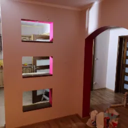 Ściana z trzema kwadratowymi otworami pomalowanymi na różowo, widok na kuchnię i korytarz z drewnianymi drzwiami, częściowo pomalowane ściany, widoczne torby i drabina.