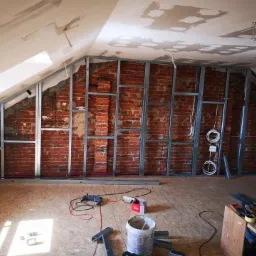 Montaż metalowej konstrukcji szkieletowej na ceglanej ścianie poddasza, widoczne narzędzia, przewody elektryczne i fragment okna.