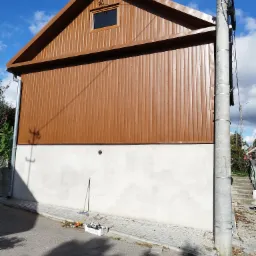 Elewacja domu jednorodzinnego z brązową, pionowo ułożoną blachą trapezową na górnej części i białą, otynkowaną dolną częścią, widok z poziomu ulicy, słup energetyczny po prawej stronie.