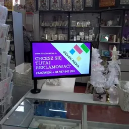 Ekran LCD wyświetlający fioletową planszę z napisem 'Chcesz się tutaj reklamować?' oraz logo firmy Gekon Media w sklepie z dewocjonaliami, w tle półki z ikonami i kartkami okolicznościowymi.