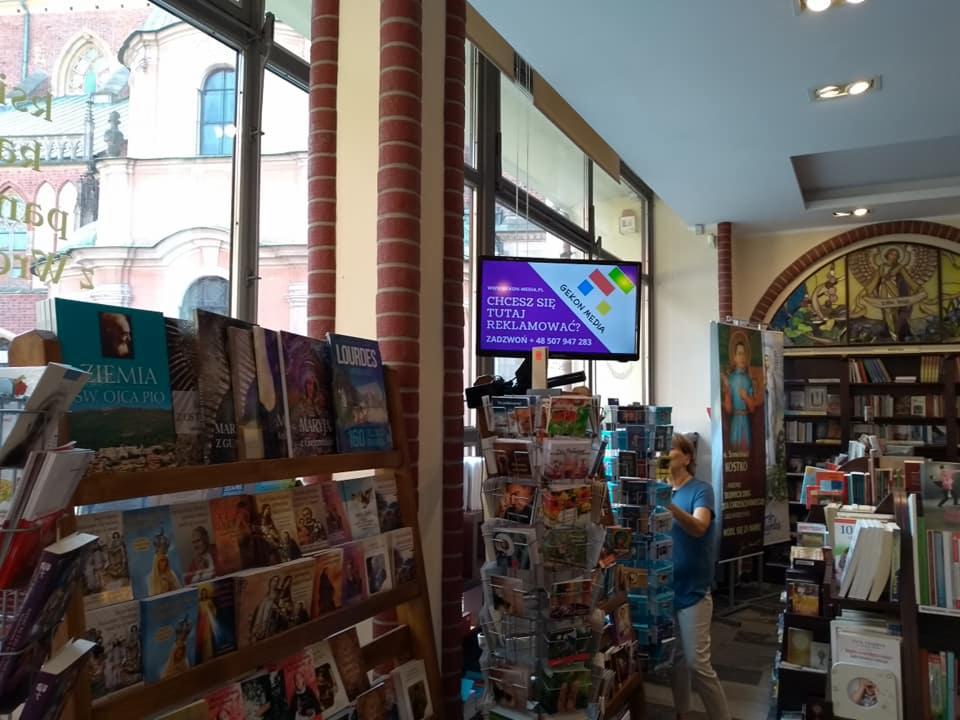Wnętrze księgarni z regałami pełnymi książek i widocznym ekranem reklamowym z napisem 'Chcesz się tutaj reklamować?' i logo Gekon Media.