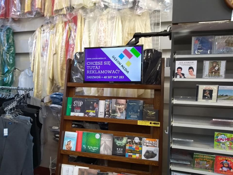 Reklama Gekon Media na ekranie w sklepie z odzieżą liturgiczną, na drewnianym regale obok książek i płyt CD.