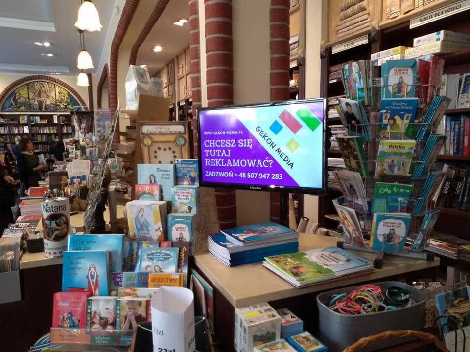 Wnętrze księgarni z ekspozycją książek dla dzieci i reklamą na ekranie LCD zachęcającą do współpracy z Gekon Media.