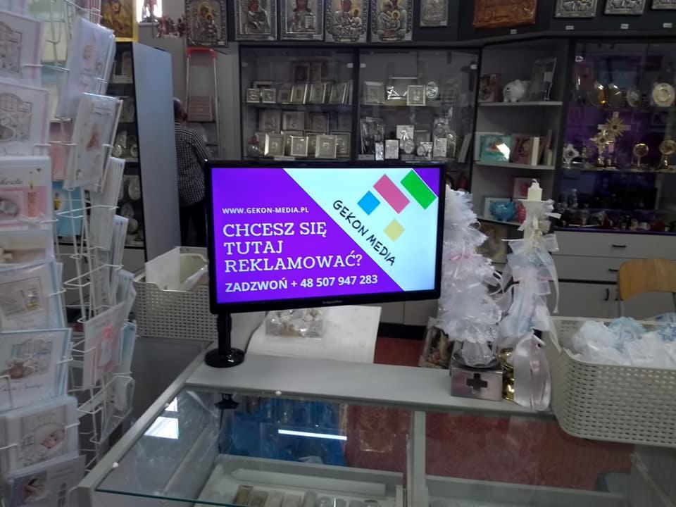 Ekran LCD wyświetlający fioletową planszę z napisem 'Chcesz się tutaj reklamować?' oraz logo firmy Gekon Media w sklepie z dewocjonaliami, w tle półki z ikonami i kartkami okolicznościowymi.