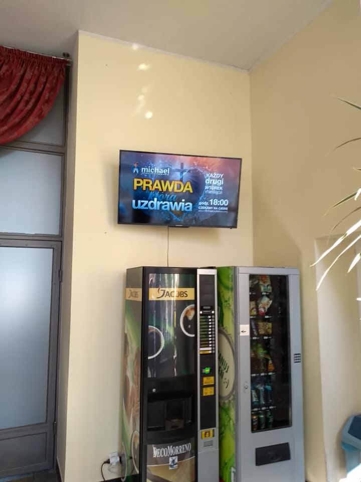 Telewizor LCD zawieszony nad dwoma automatami z napojami i przekąskami w jasnym pomieszczeniu z zasłoniętym oknem po lewej stronie i rośliną po prawej; na ekranie telewizora wyświetlana jest...