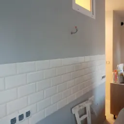 Ściana z ułożonymi białymi, błyszczącymi płytkami typu metro, z widocznymi puszkami elektrycznymi, pomalowana na szaro powyżej, z małym oknem i krzesłem malarskim w tle.