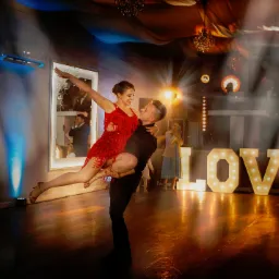 Para taneczna w Siedlcach w dynamicznej pozie na tle dekoracji świetlnych i napisu LOVE, kobieta w czerwonej sukience z frędzlami, mężczyzna w czarnym stroju.