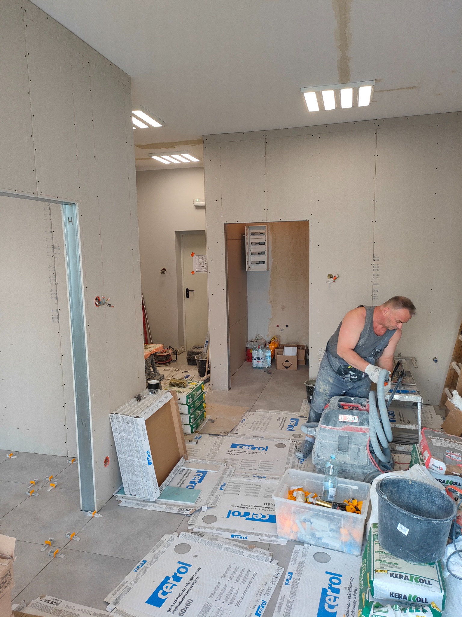 Pracownik w szarym kombinezonie tnie płytki na stole roboczym w trakcie remontu pomieszczenia z płytami kartonowo-gipsowymi na ścianach i częściowo ułożonymi płytkami podłogowymi z użyciem...