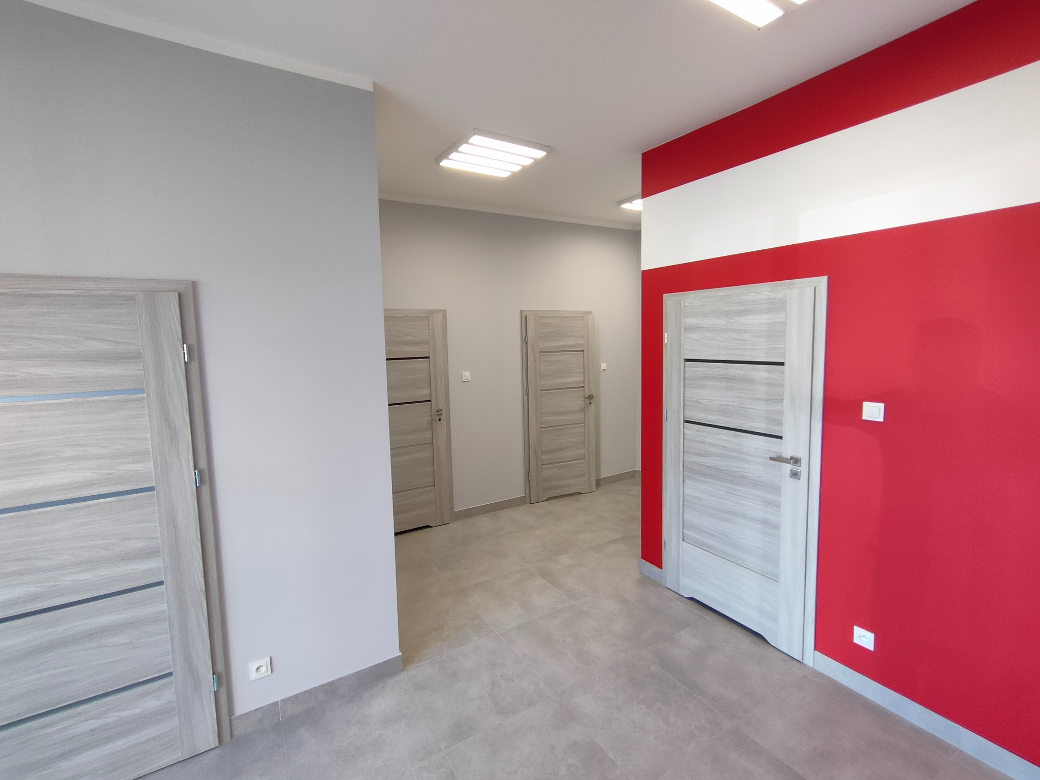 Wnętrze biura po remoncie: szare ściany, szara podłoga, drzwi w kolorze jasnego drewna z poziomymi paskami, ściana pomalowana na czerwono z białym pasem u góry.