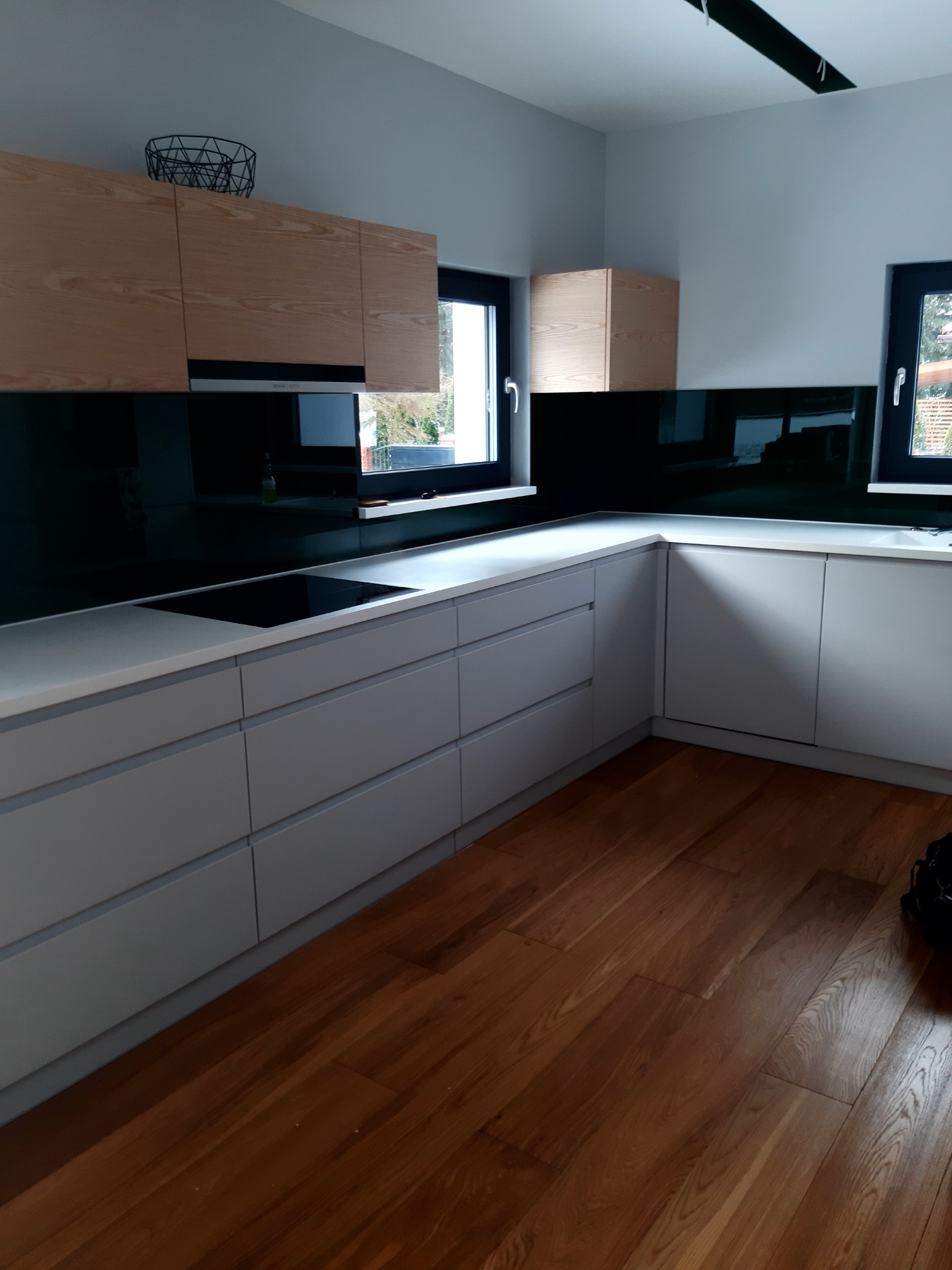 Nowoczesna kuchnia w Krzeszowicach z drewnianymi szafkami górnymi i szklanym panelem między blatem a szafkami, odbijającym światło z okna.