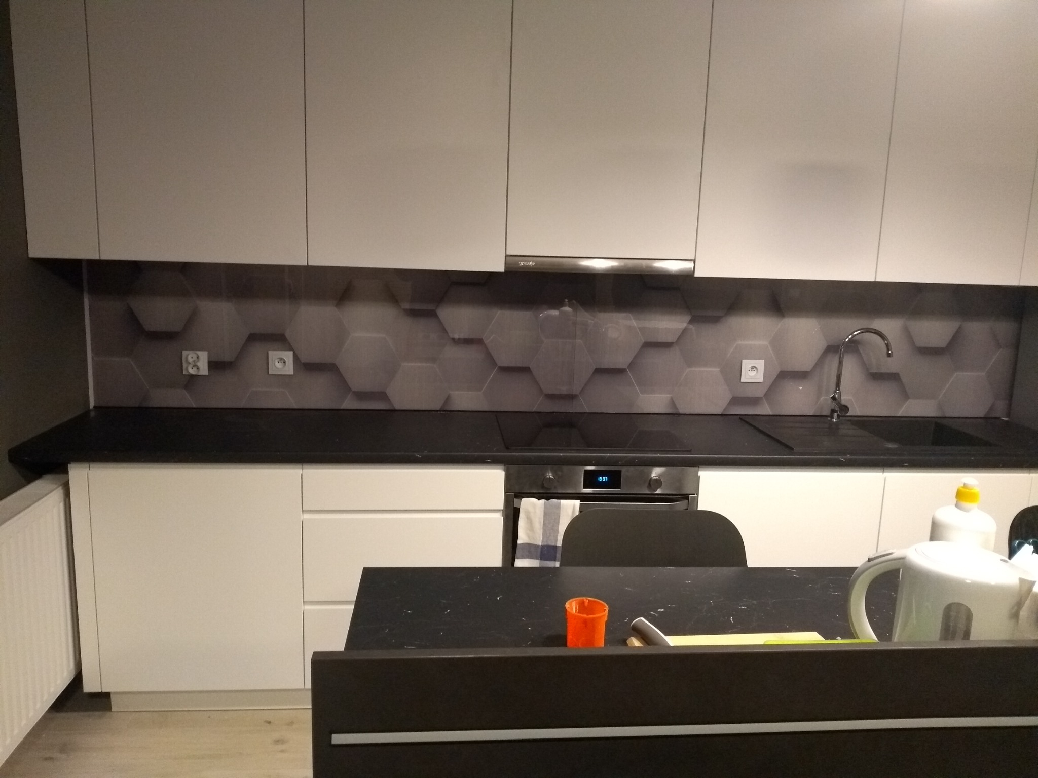 Nowoczesna, minimalistyczna kuchnia z białymi szafkami, czarnym blatem i szarą mozaiką heksagonalną nad blatem. Widoczny czajnik elektryczny i inne akcesoria kuchenne.