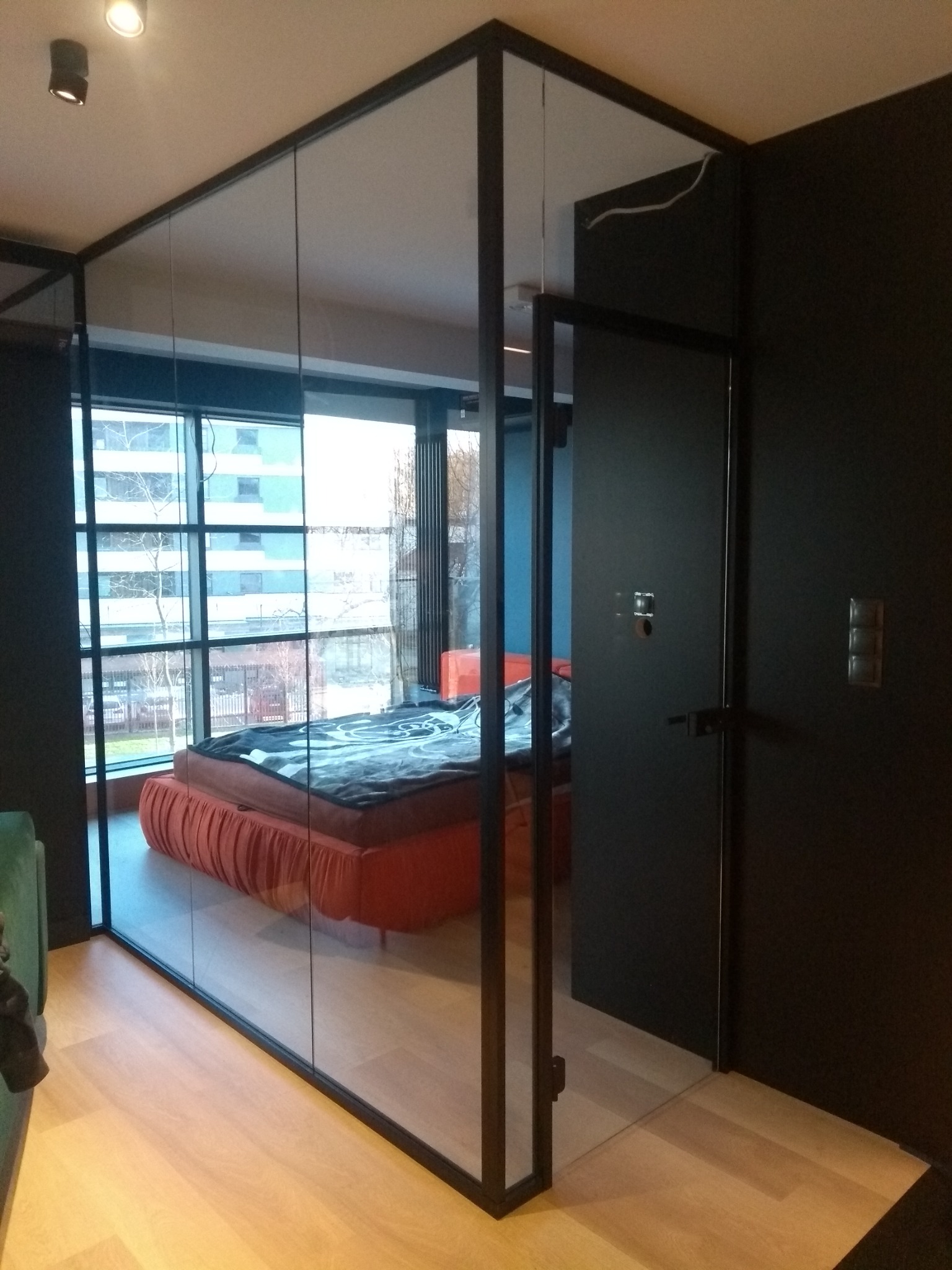 Szklana ściana działowa z czarną ramą oddzielająca sypialnię, widok na łóżko z pomarańczową ramą i dużą szybą okienną w tle.