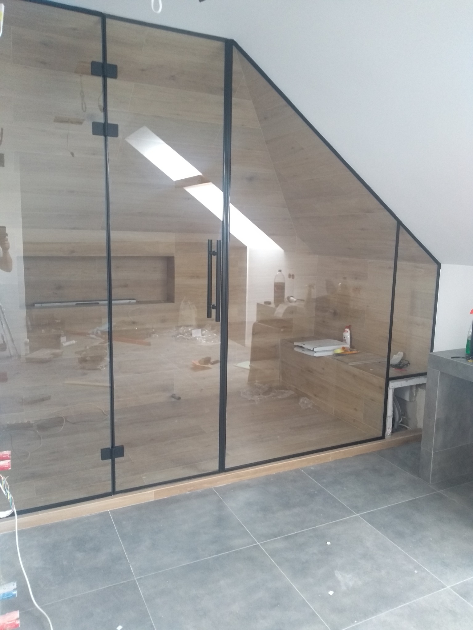 Szklana ścianka działowa z czarnymi profilami w łazience na poddaszu, widoczne płytki imitujące drewno na ścianach i szare płytki na podłodze, stan surowy.