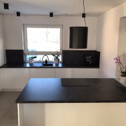 Stolmeblux - Nowoczesna kuchnia z białą zabudową i czarnym blatem. Na wyspie płyta indukcyjna, a w oknie doniczki z roślinami. Minimalistyczny design z wiszącą lampą.