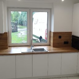 Stolmeblux - Nowoczesna, biała kuchnia z drewnianym blatem i oknem, przez które widać dostawczy samochód. Minimalistyczny design, fronty na wysoki połysk.