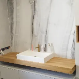 Nowoczesna łazienka z blatem z drewna i szafką w kolorze szarym, białą umywalką nablatową i marmurową ścianą. Złota bateria i akcesoria łazienkowe.