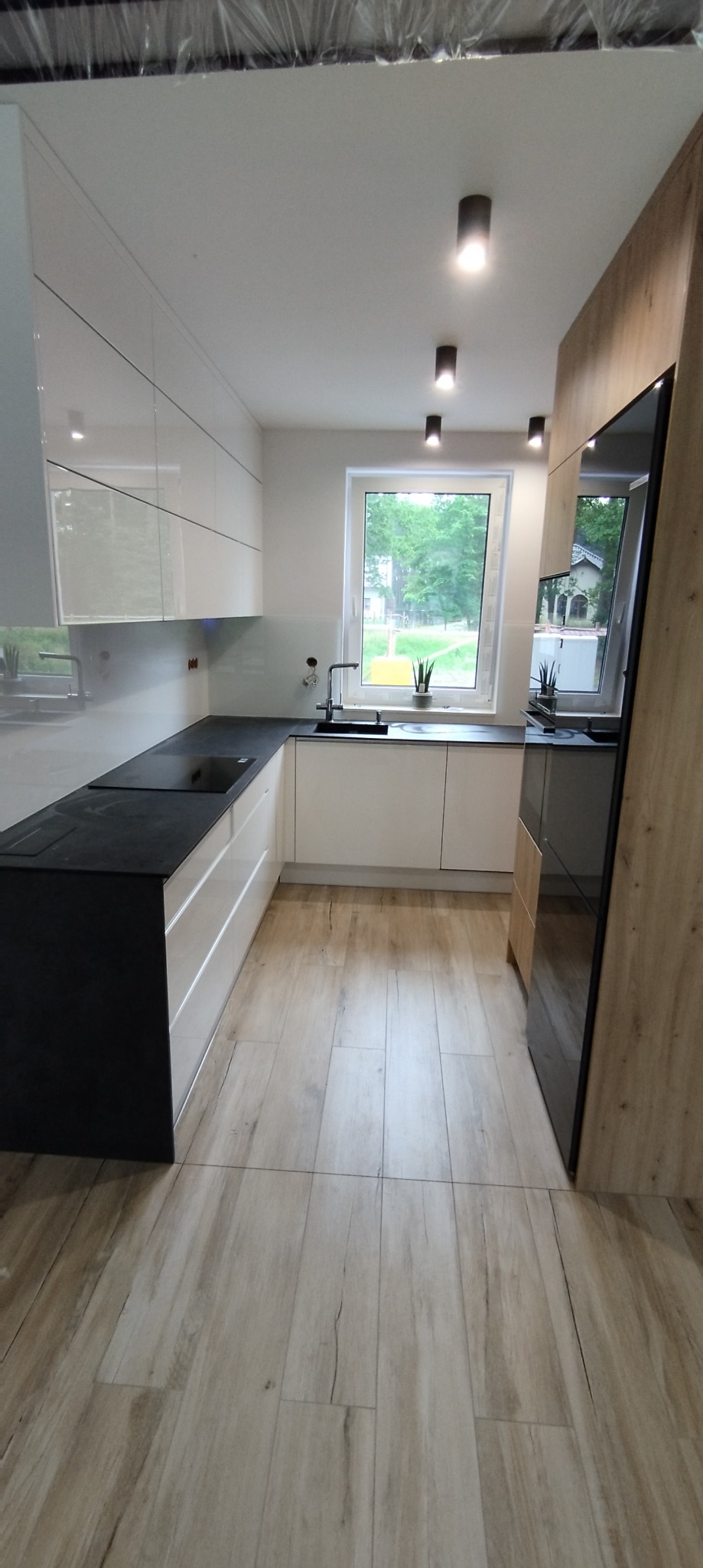 Nowoczesna kuchnia z białymi, błyszczącymi szafkami, czarnym blatem i drewnopodobną podłogą z płytek, oświetlona punktowymi lampami sufitowymi, z widokiem na zieleń za oknem.