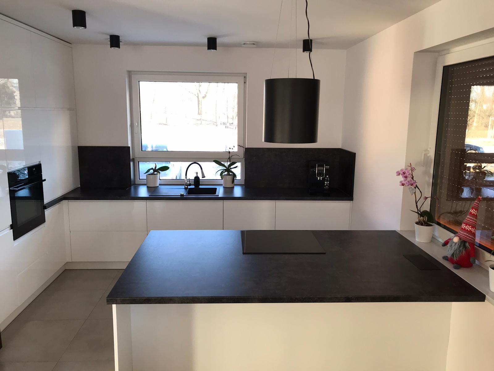 Nowoczesna kuchnia z białą zabudową i czarnym blatem. Na wyspie płyta indukcyjna, a w oknie doniczki z roślinami. Minimalistyczny design z wiszącą lampą.