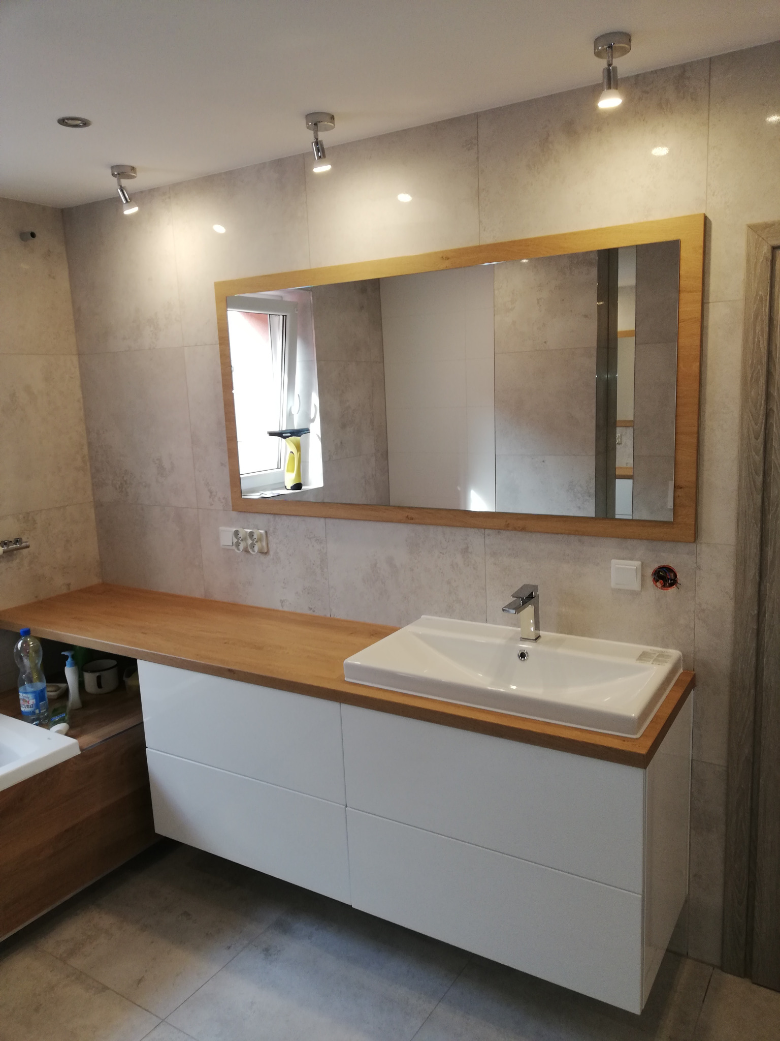 Nowoczesna łazienka z drewnianym blatem, białą szafką pod umywalkę i dużym lustrem w drewnianej ramie. Jasne płytki na ścianach, punktowe oświetlenie sufitowe.