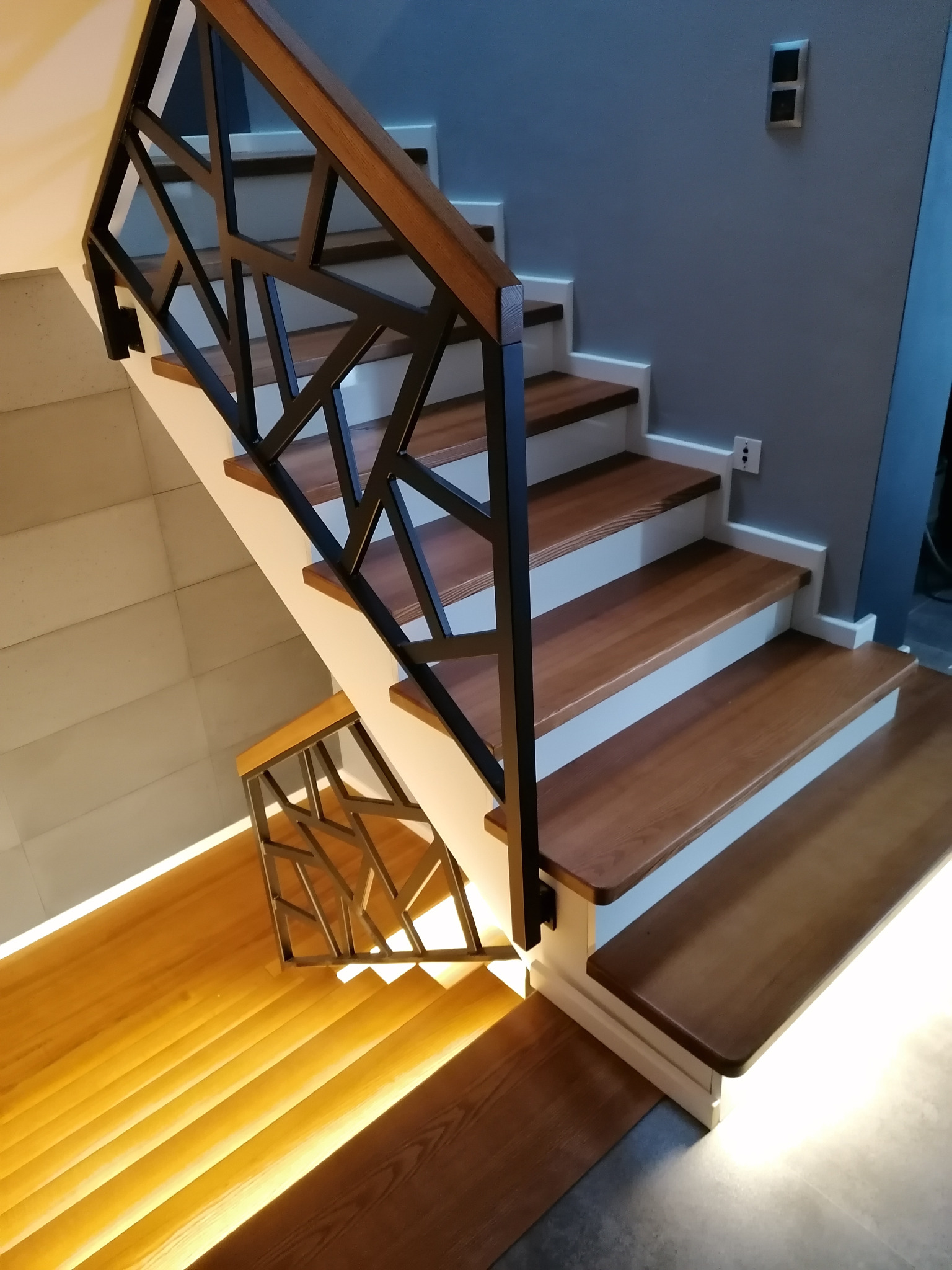 Nowoczesne schody drewniane z geometryczną, czarną balustradą i podświetleniem LED stopni. Minimalistyczny design, drewno o ciepłym odcieniu i szara ściana w tle.
