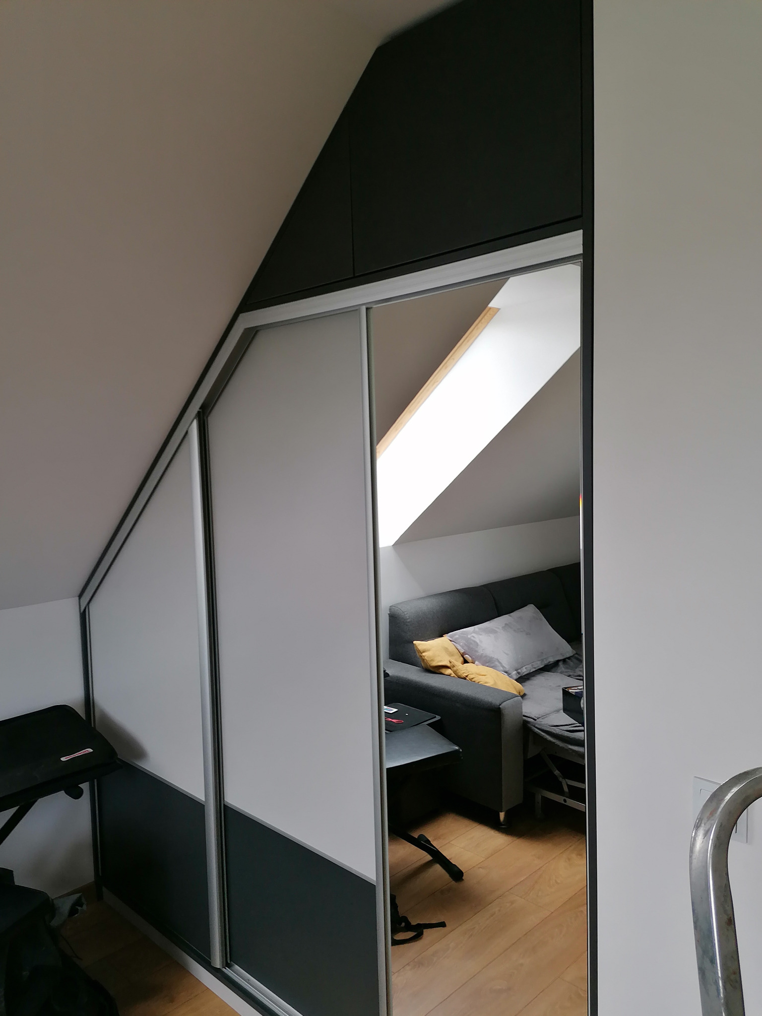 Dwukolorowa szafa wnękowa z przesuwnymi drzwiami w pokoju na poddaszu, widok na wnętrze pokoju z szarą sofą i oknem dachowym.