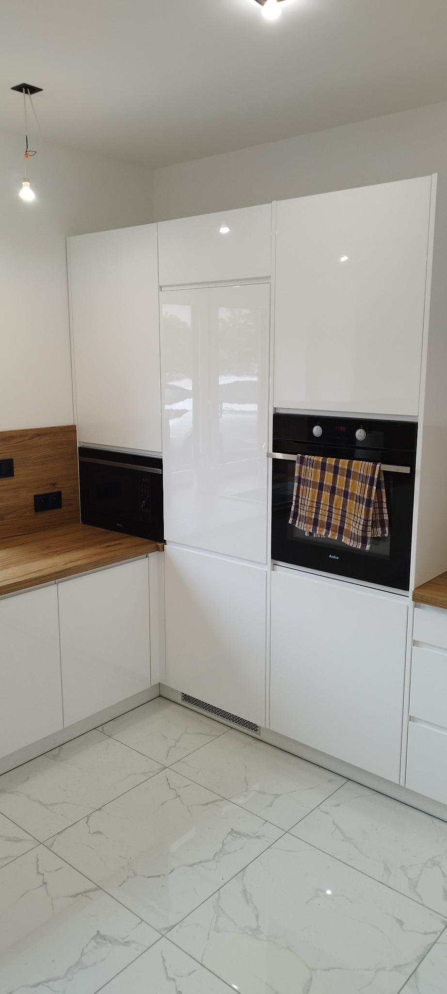 Biała, nowoczesna kuchnia z drewnianym blatem i zabudowanymi urządzeniami AGD. Widoczne płytki podłogowe imitujące marmur i minimalistyczne oświetlenie.