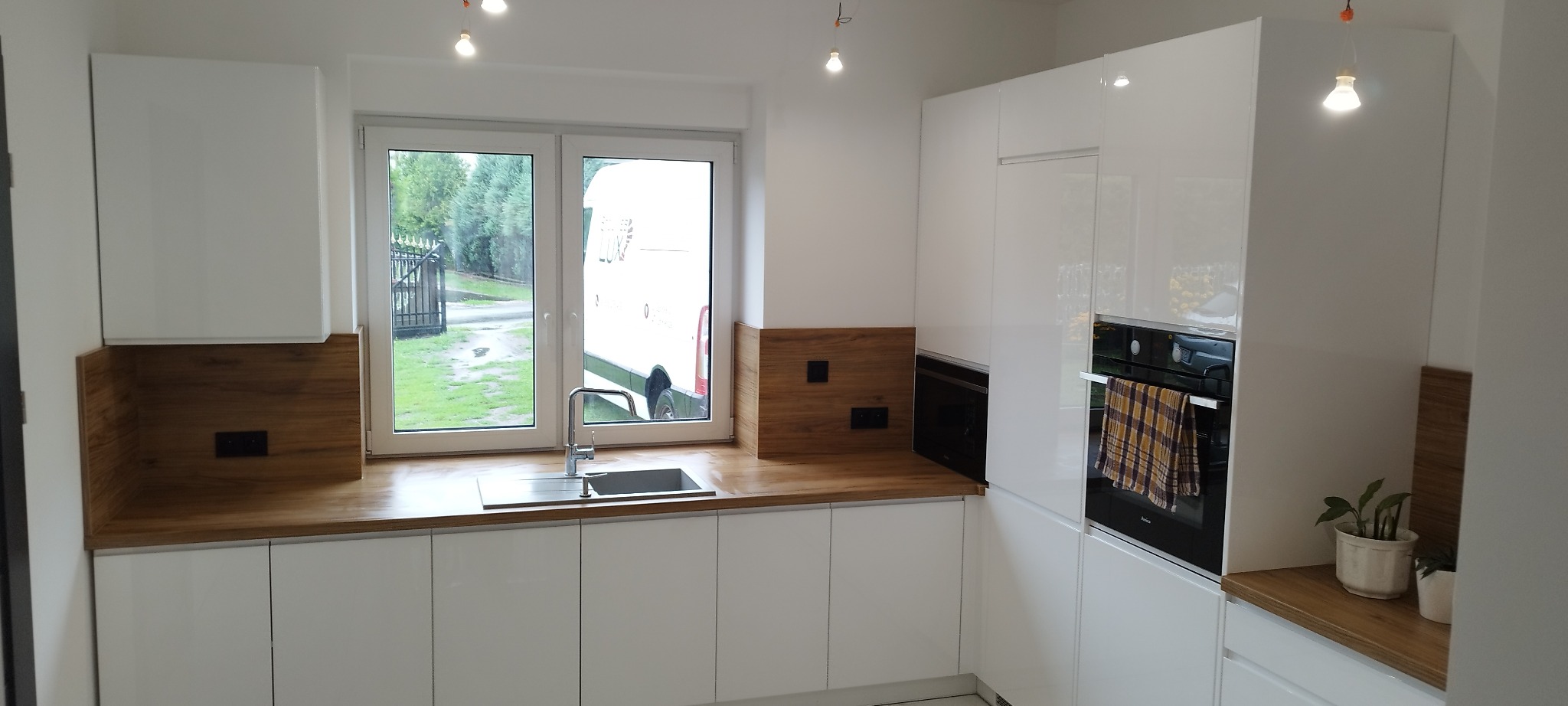 Nowoczesna, biała kuchnia z drewnianymi akcentami i oknem, przez które widać samochód dostawczy. Minimalistyczny design z piekarnikiem i rośliną doniczkową.