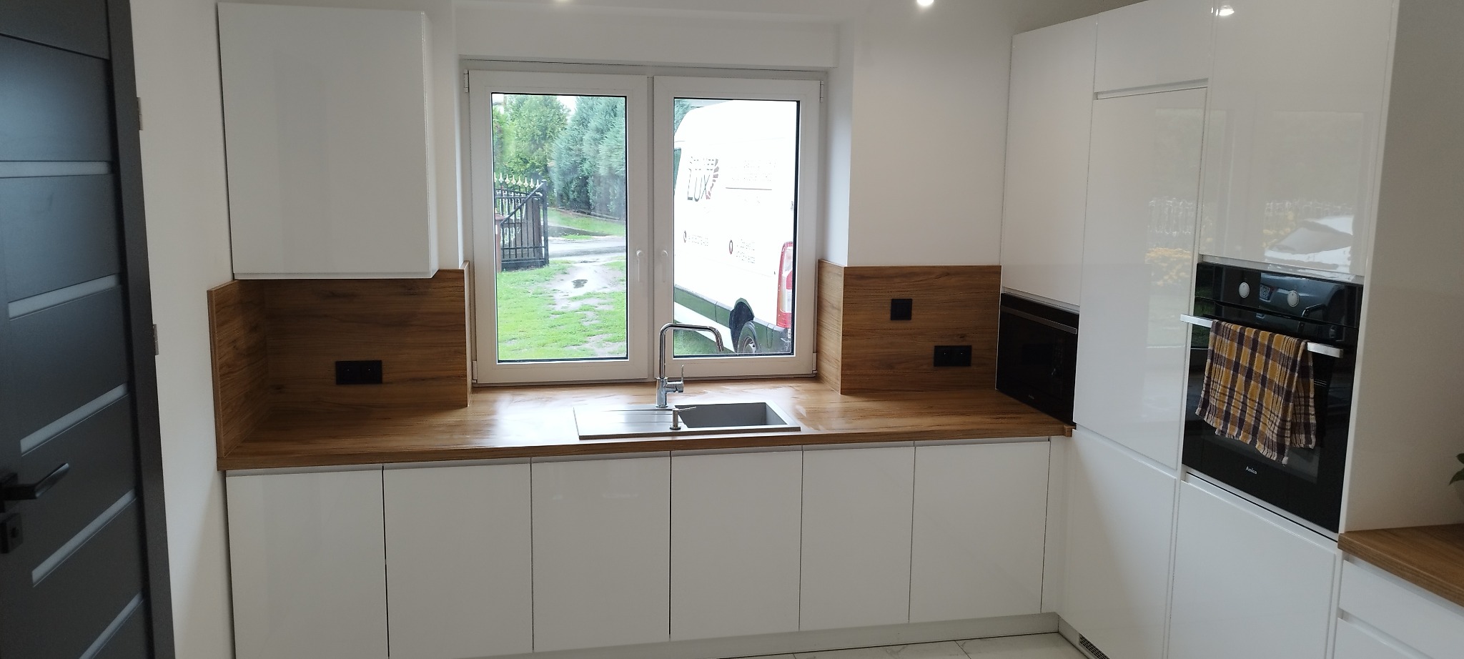 Nowoczesna, biała kuchnia z drewnianym blatem i oknem, przez które widać dostawczy samochód. Minimalistyczny design, fronty na wysoki połysk.