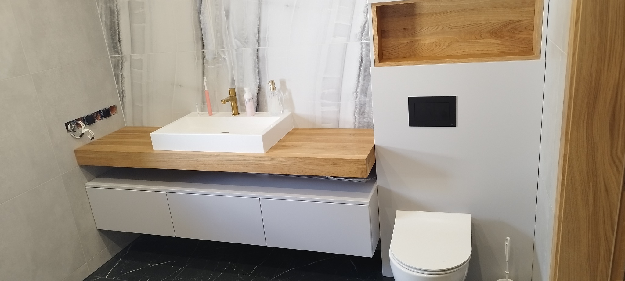 Nowoczesna łazienka z drewnianym blatem pod umywalką nablatową, szarą szafką podwieszaną i marmurowymi płytkami na ścianie. Widoczna toaleta i drewniana półka.