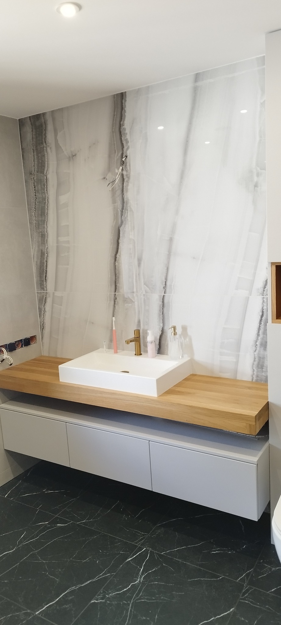 Nowoczesna łazienka z blatem z drewna i szafką w kolorze szarym, białą umywalką nablatową i marmurową ścianą. Złota bateria i akcesoria łazienkowe.