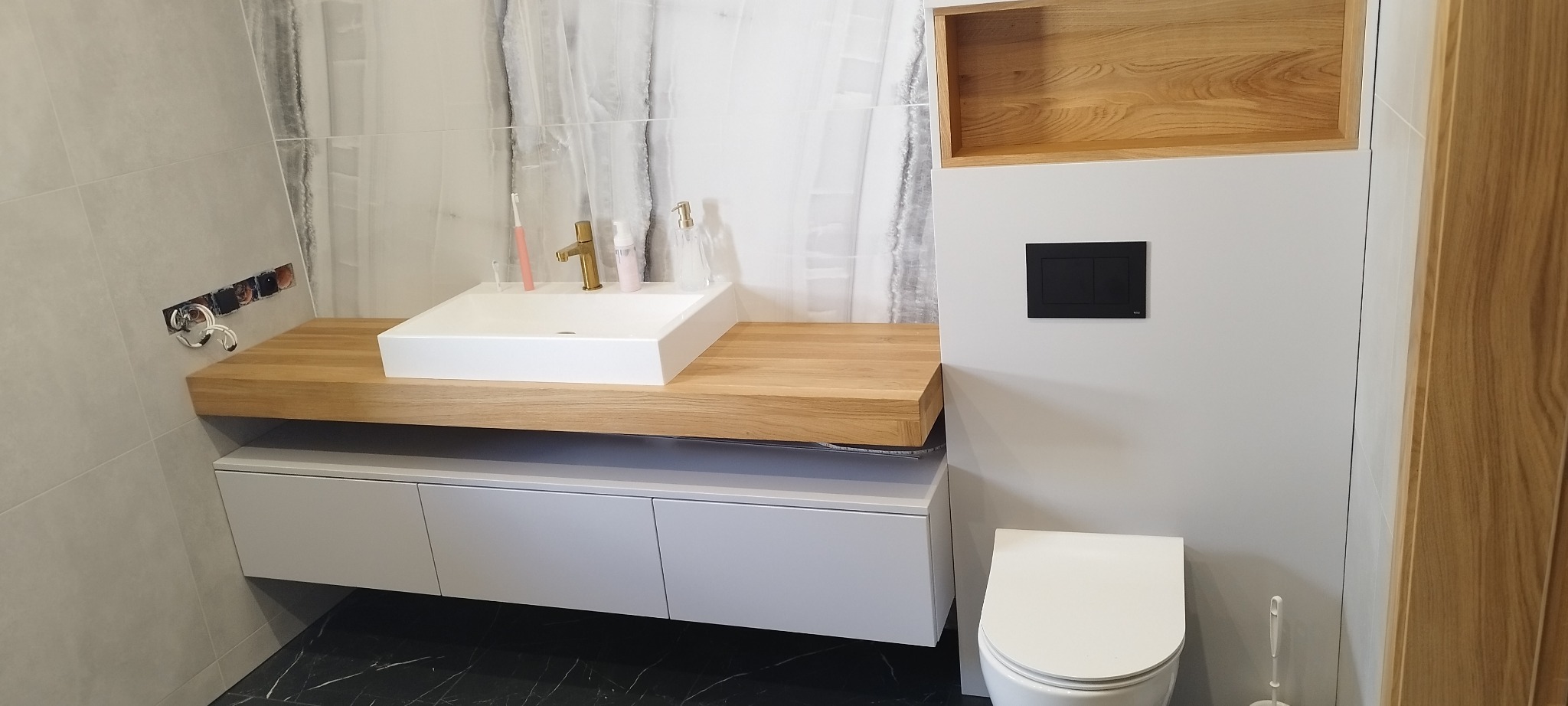 Nowoczesna łazienka: biała umywalka nablatowa na drewnianym blacie, szare szafki pod umywalką, wisząca toaleta i drewniana wnęka nad nią, marmurowe płytki na ścianie.